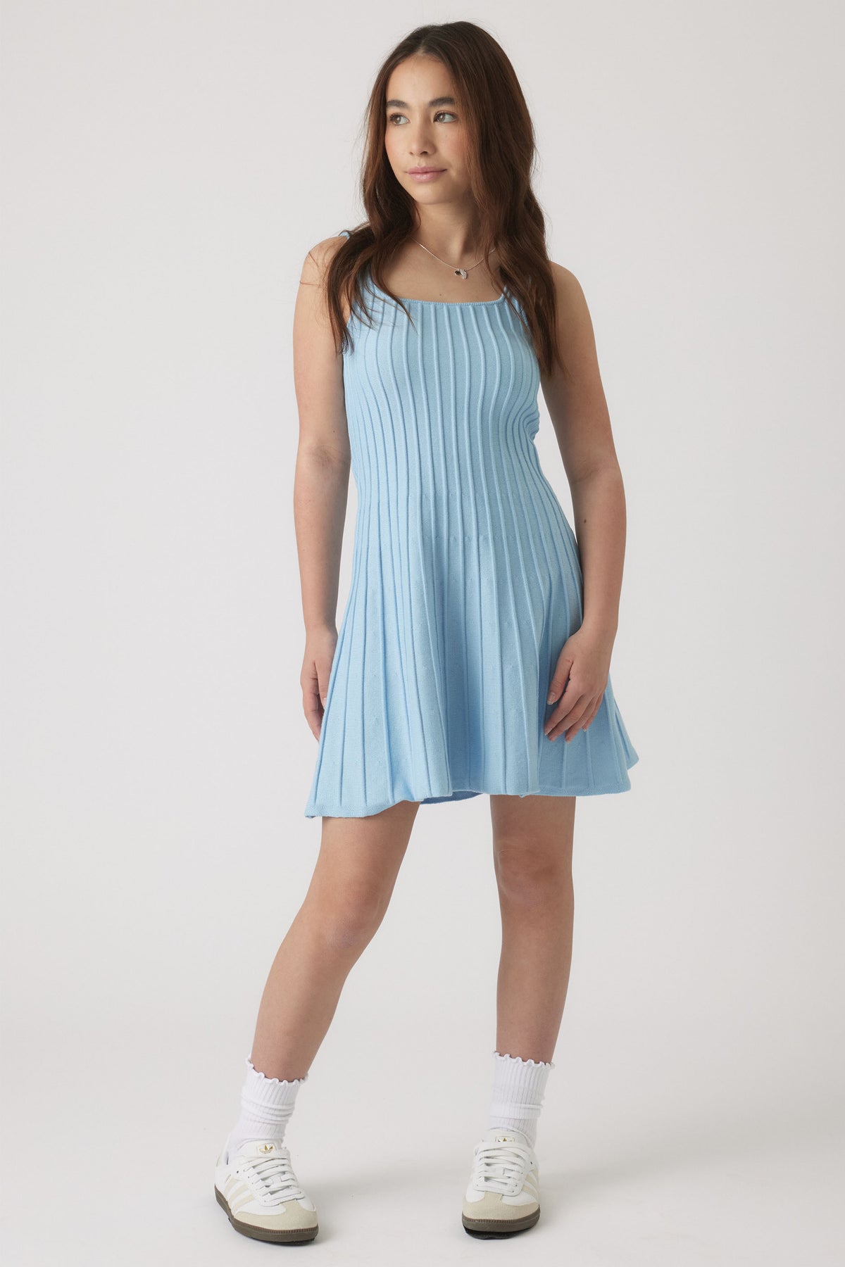 Girl wearing a light blue knit dress with vertical ribbing and a flared hem, styled with white socks and sneakers.