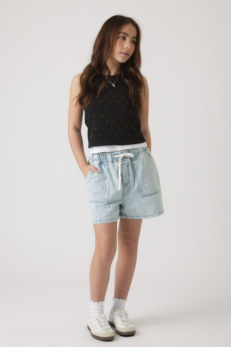 Girl wearing a black crystal-embellished tank paired with light denim shorts, white socks, and sneakers in a casual pose.