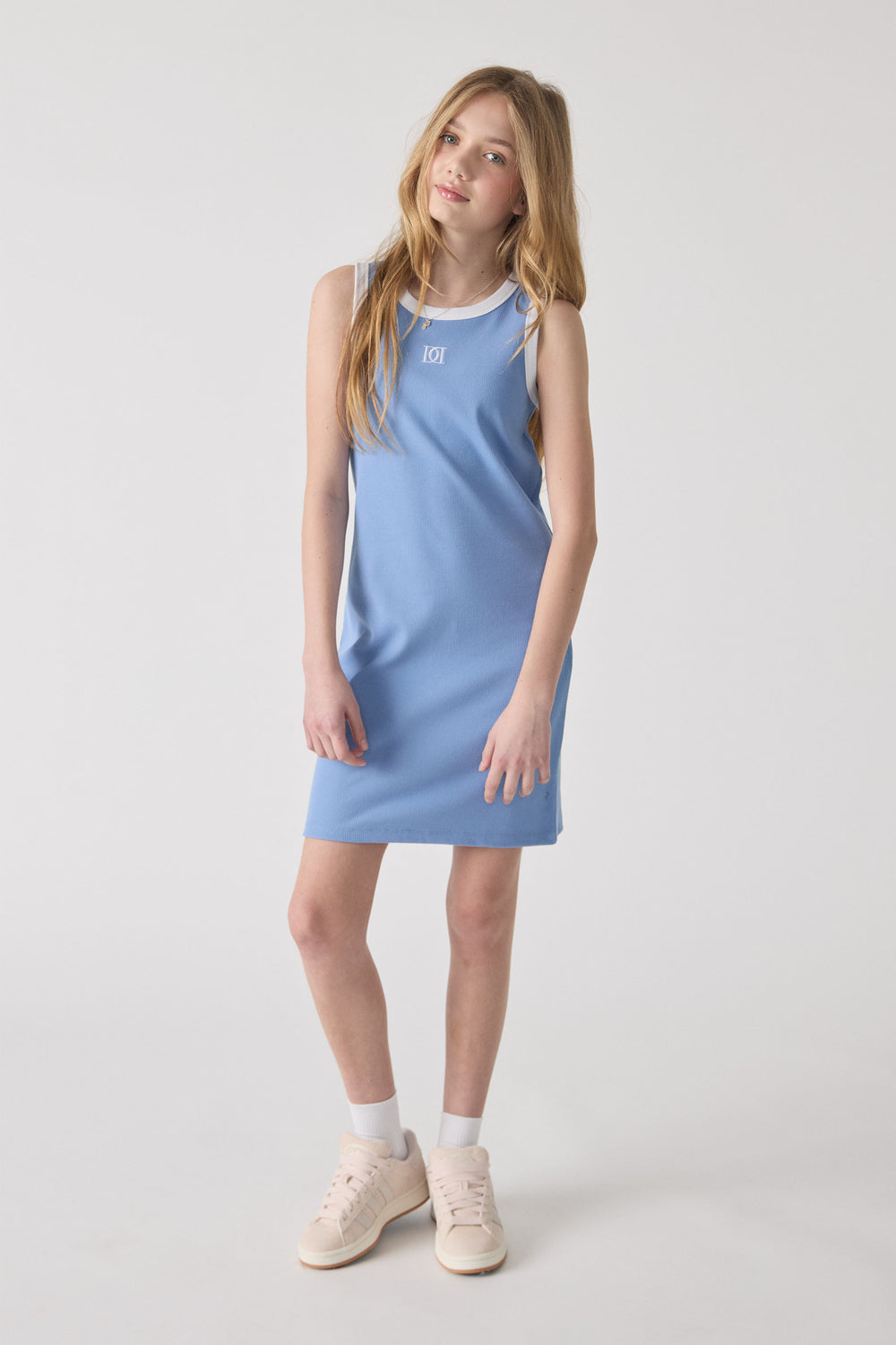 Young girl wearing a sleeveless dusty blue monogram dress with white trim, styled with white socks and cream sneakers.
