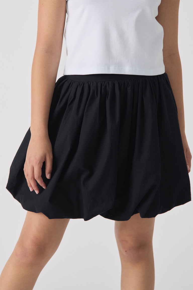 Girl wearing a black bubble mini skirt with gathered waist paired with a white sleeveless top, shown from front.