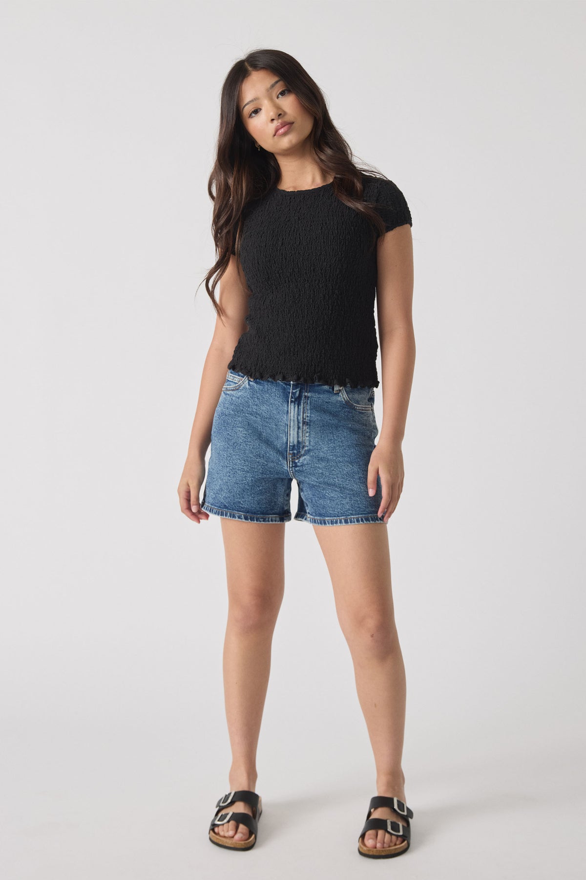 Teen girl wearing relaxed fit mid-blue denim shorts paired with a textured black short sleeve top and black slide sandals