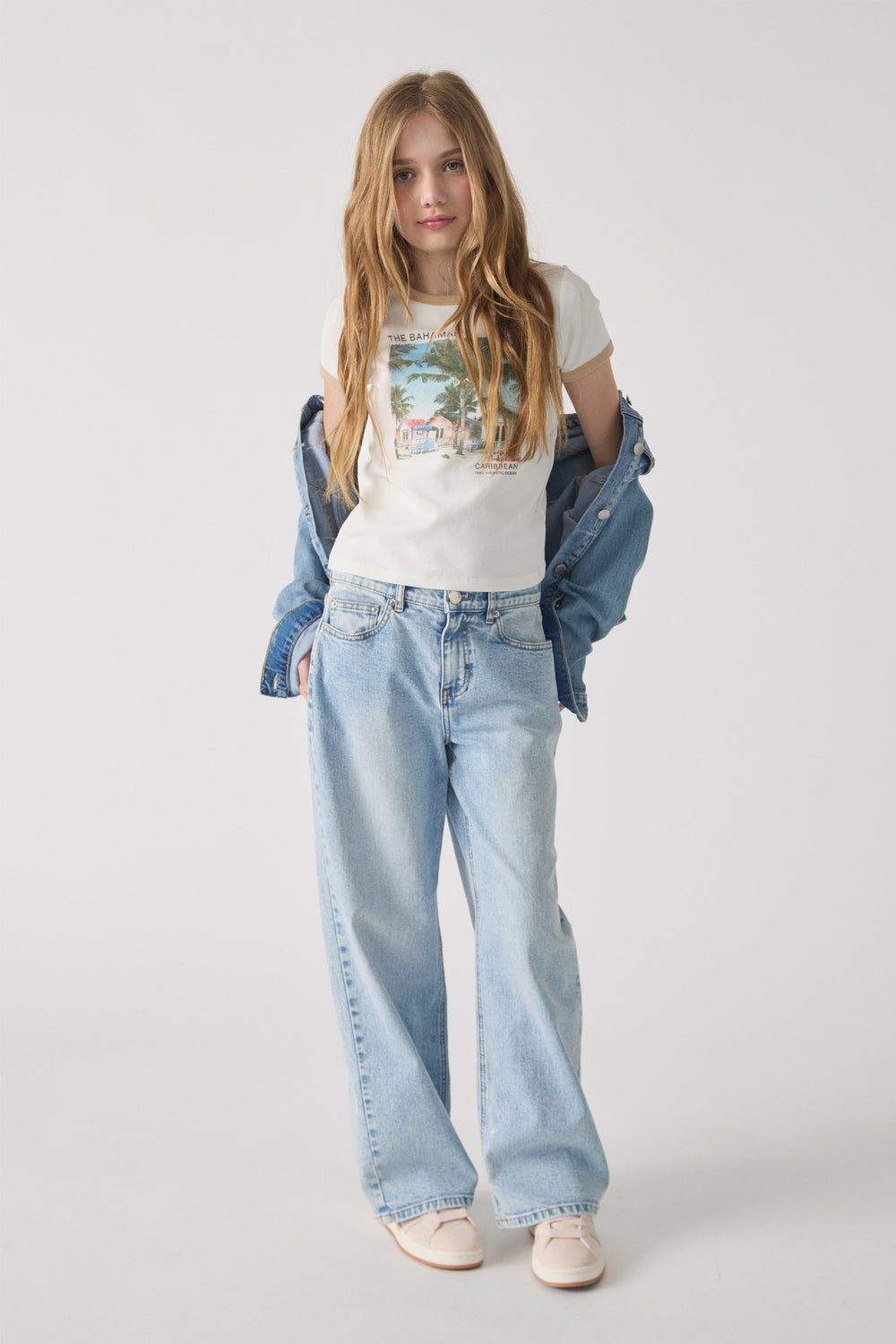 Teen girl wearing a vintage graphic crop tee paired with relaxed light wash wide-leg denim jeans and a denim jacket draped at elbows