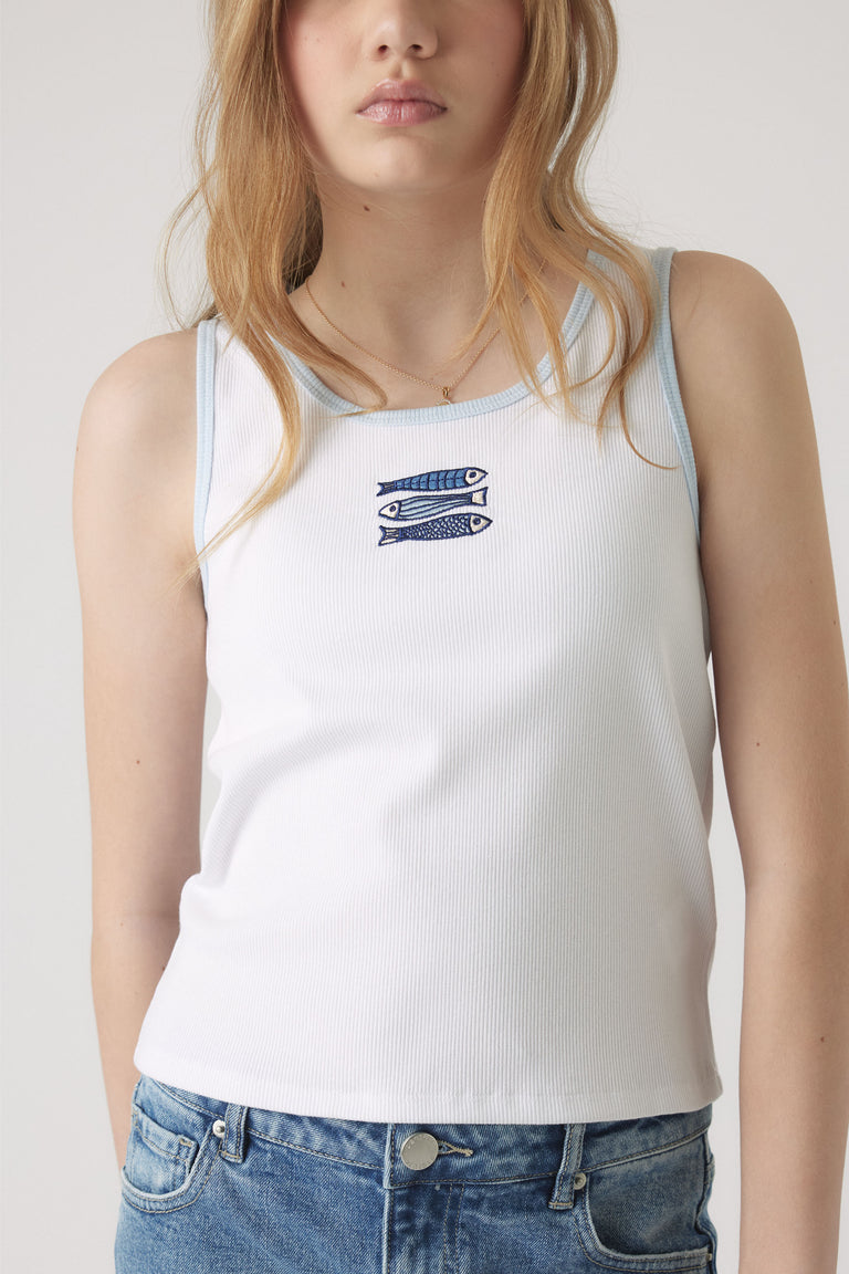 Girl wearing a white ribbed tank with blue trim and a small embroidered fish design, paired with light blue denim jeans.