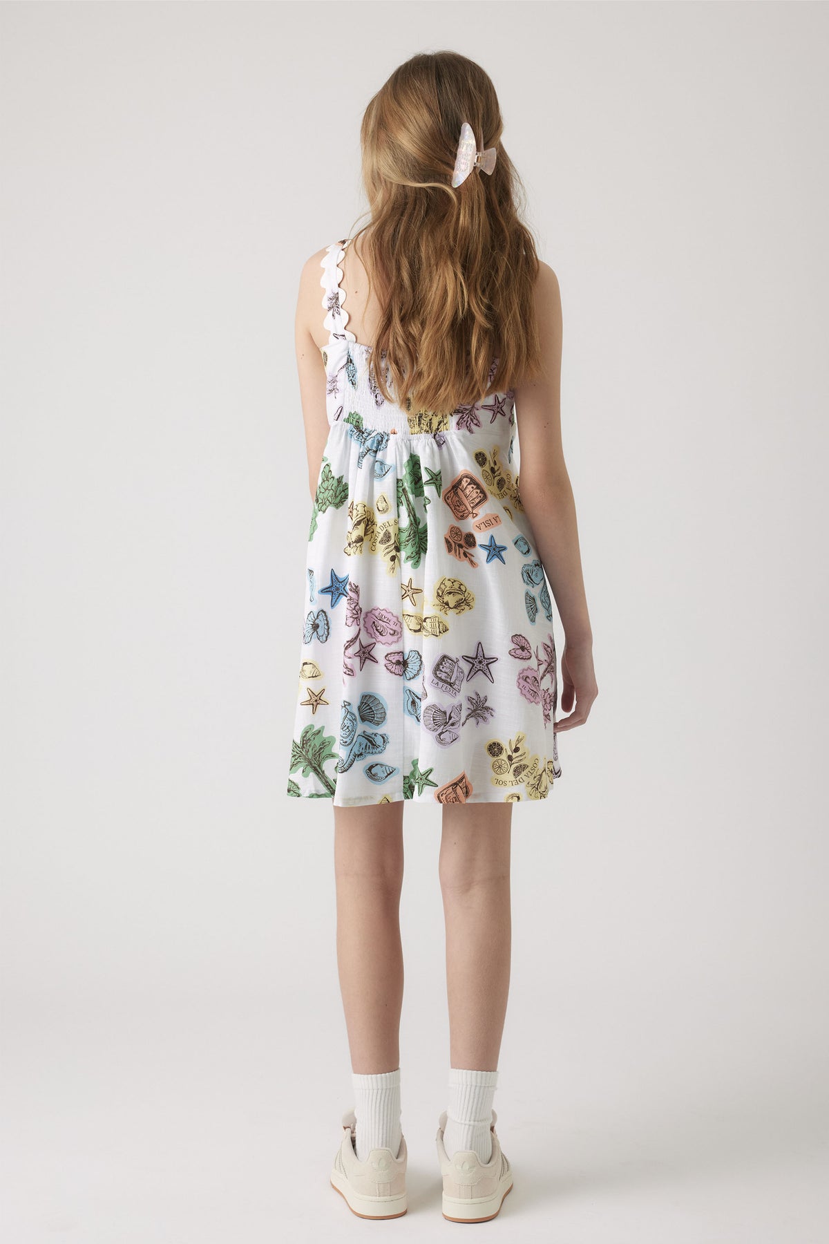 Girl wearing a white strappy dress with colorful sea creature prints, paired with white socks and sneakers, hair clipped back.