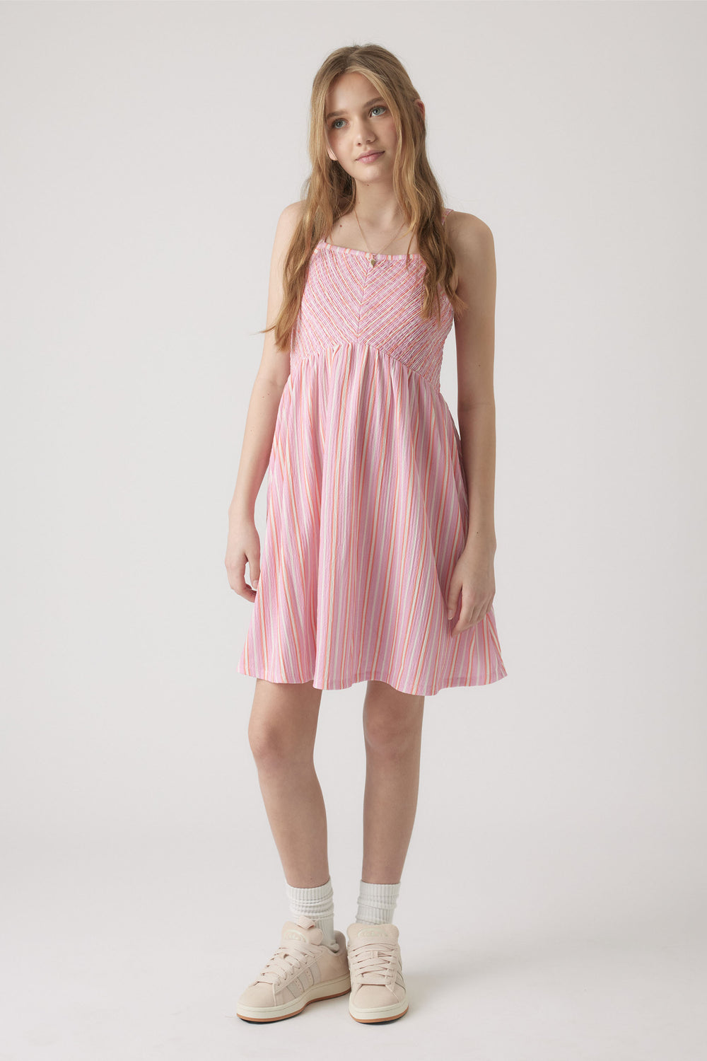 Girl wearing a pink and white striped shirred dress with spaghetti straps, paired with white socks and sneakers.