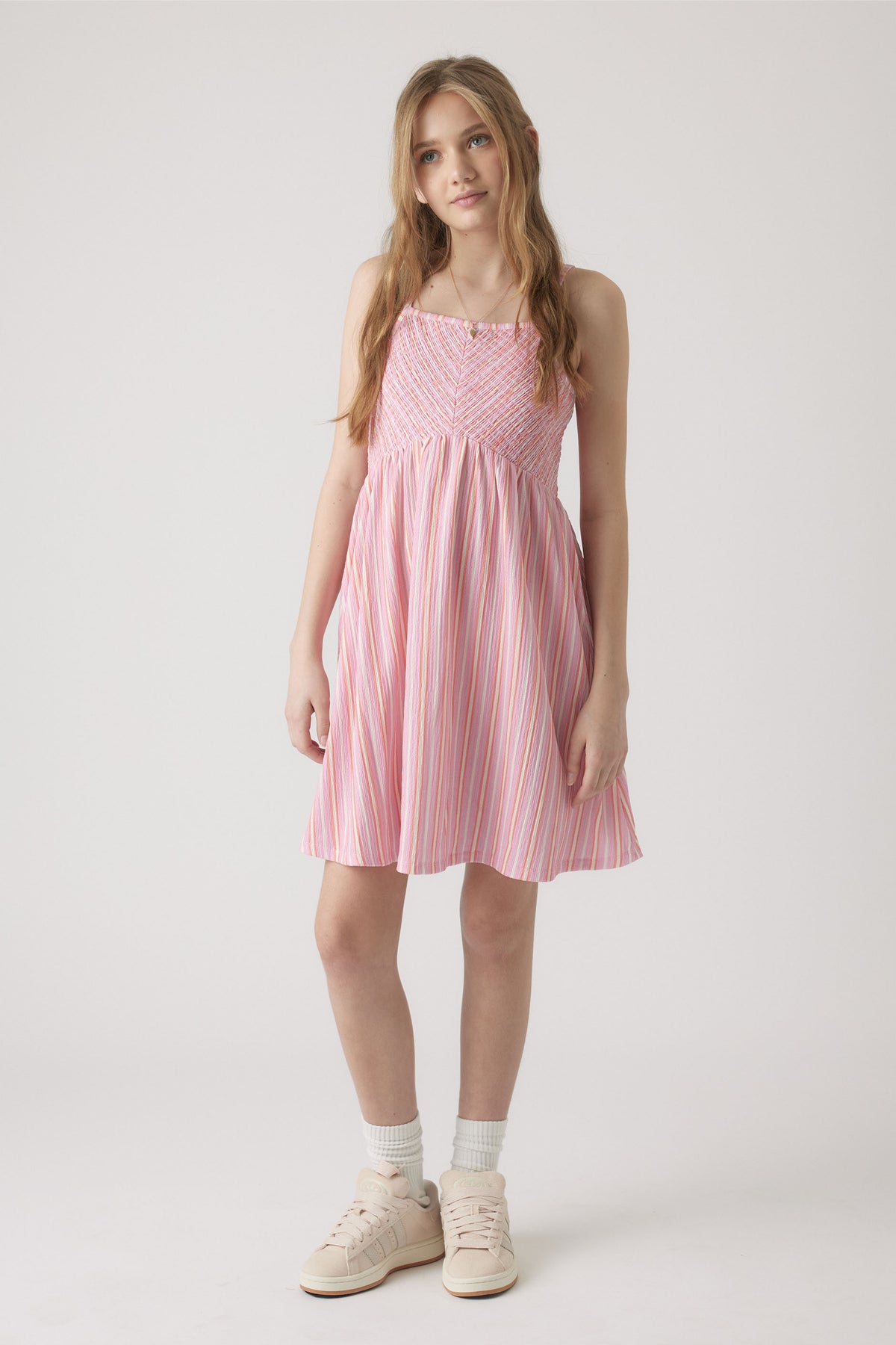 Girl wearing a pink and white striped shirred dress with spaghetti straps, paired with white socks and sneakers.