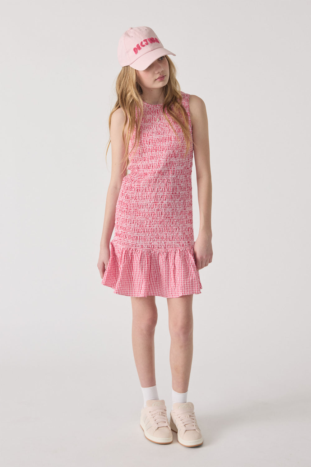 Girl wearing a sleeveless pink gingham dress with smocked bodice and ruffle hem paired with a pink cap and sneakers