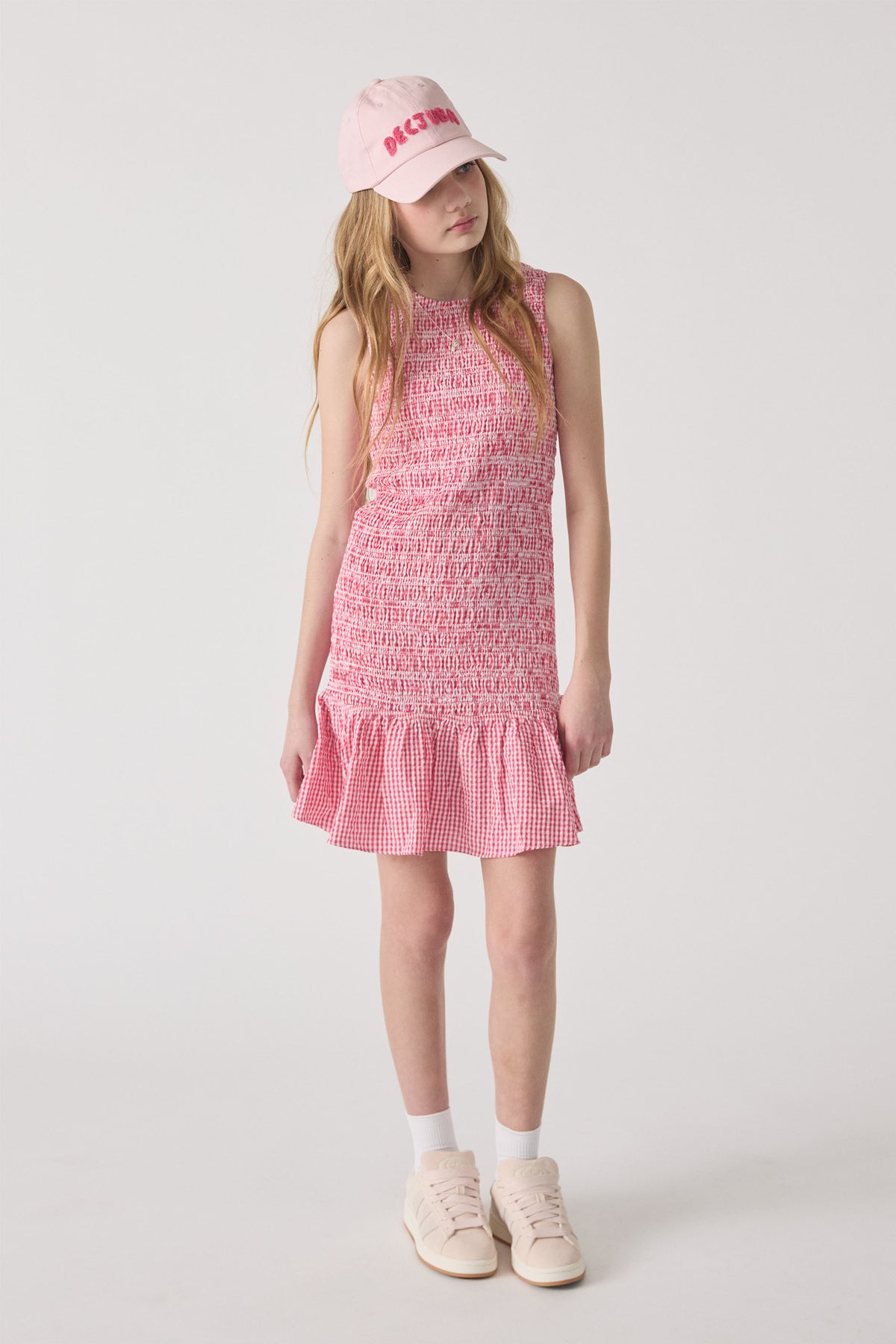 Girl wearing a sleeveless pink gingham dress with smocked bodice and ruffle hem paired with a pink cap and sneakers