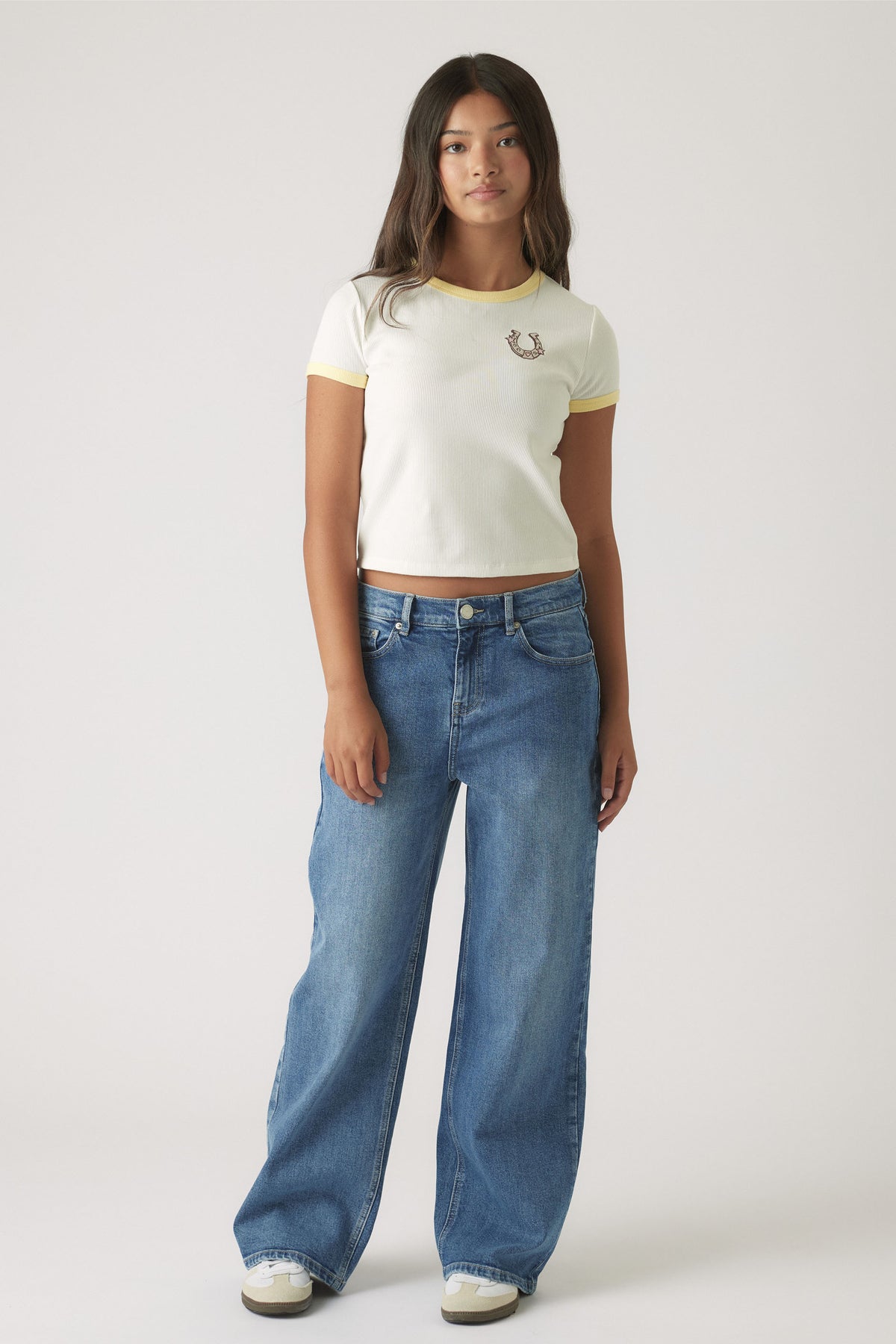 Teen girl wearing a white cropped ringer tee with yellow trim paired with relaxed wide-leg blue denim jeans and sneakers
