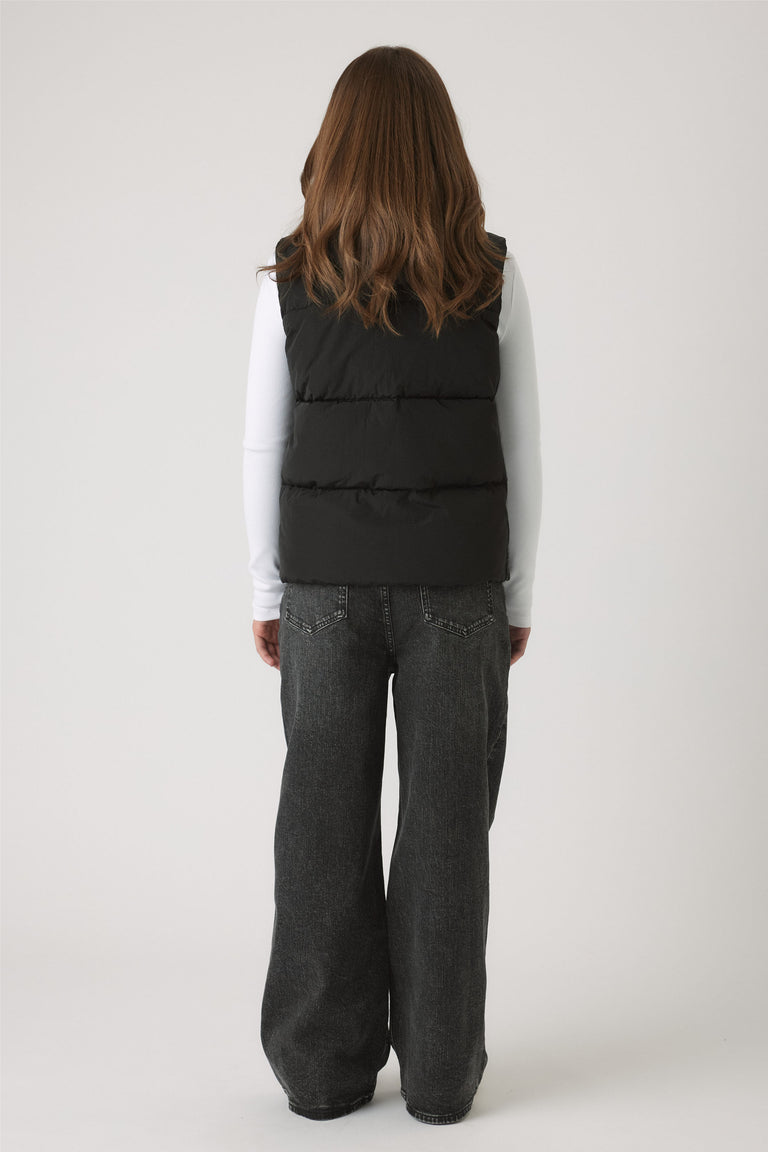 Back view of Girls Hooded Puffer Vest in black worn over white long sleeves and loose dark denim jeans by DECJUBA.