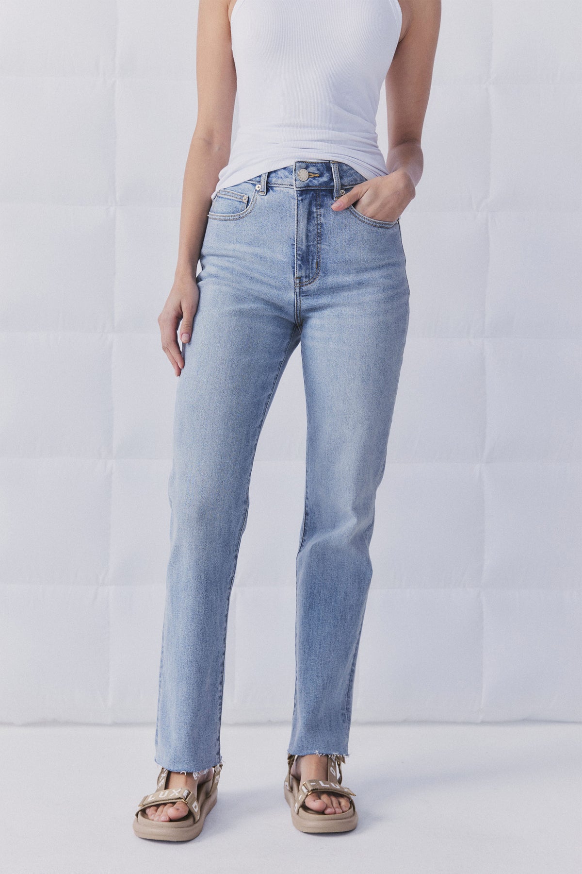 Woman wearing light blue straight leg jeans with a raw hem paired with a white sleeveless top and beige sandals