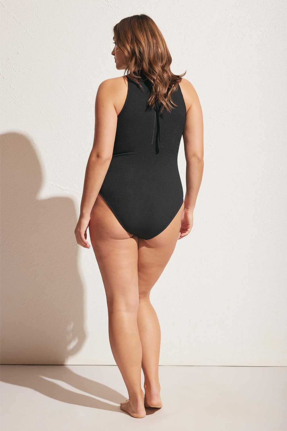 Woman wearing a black crinkle racer one piece with a high neckline and tie detail, standing barefoot against a wall