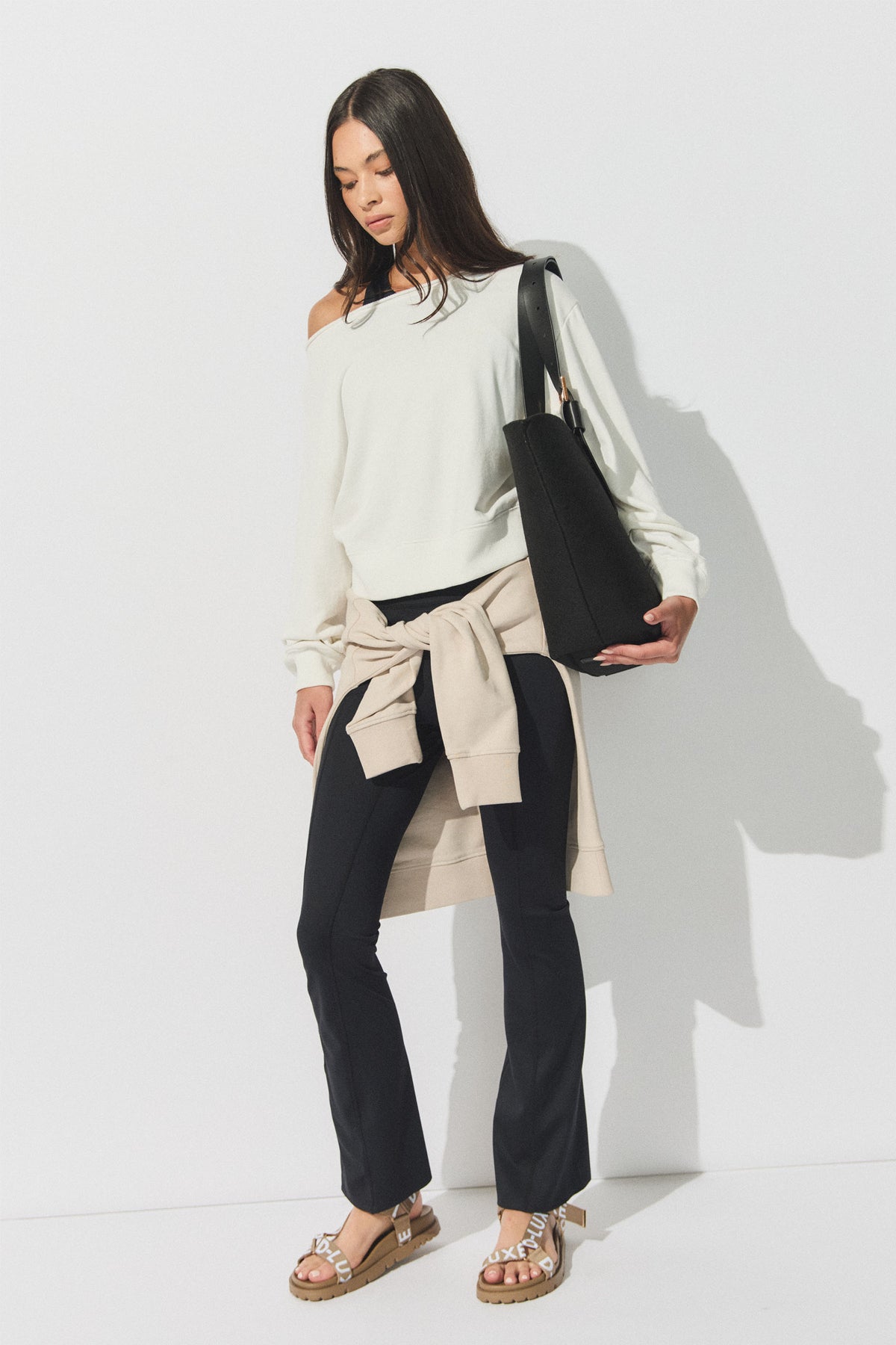 Woman wearing black flare leggings with a cream off-shoulder sweater, beige sweatshirt tied at waist, and sandals, holding a black tote bag.