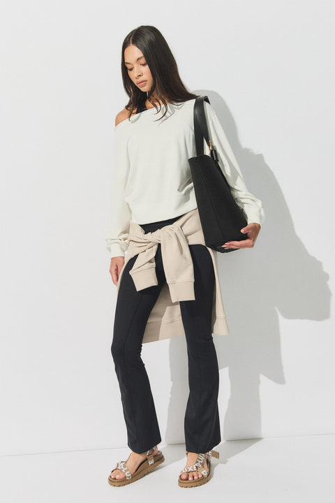 Woman wearing black flare leggings with a cream off-shoulder sweater, beige sweatshirt tied at waist, and sandals, holding a black tote bag.