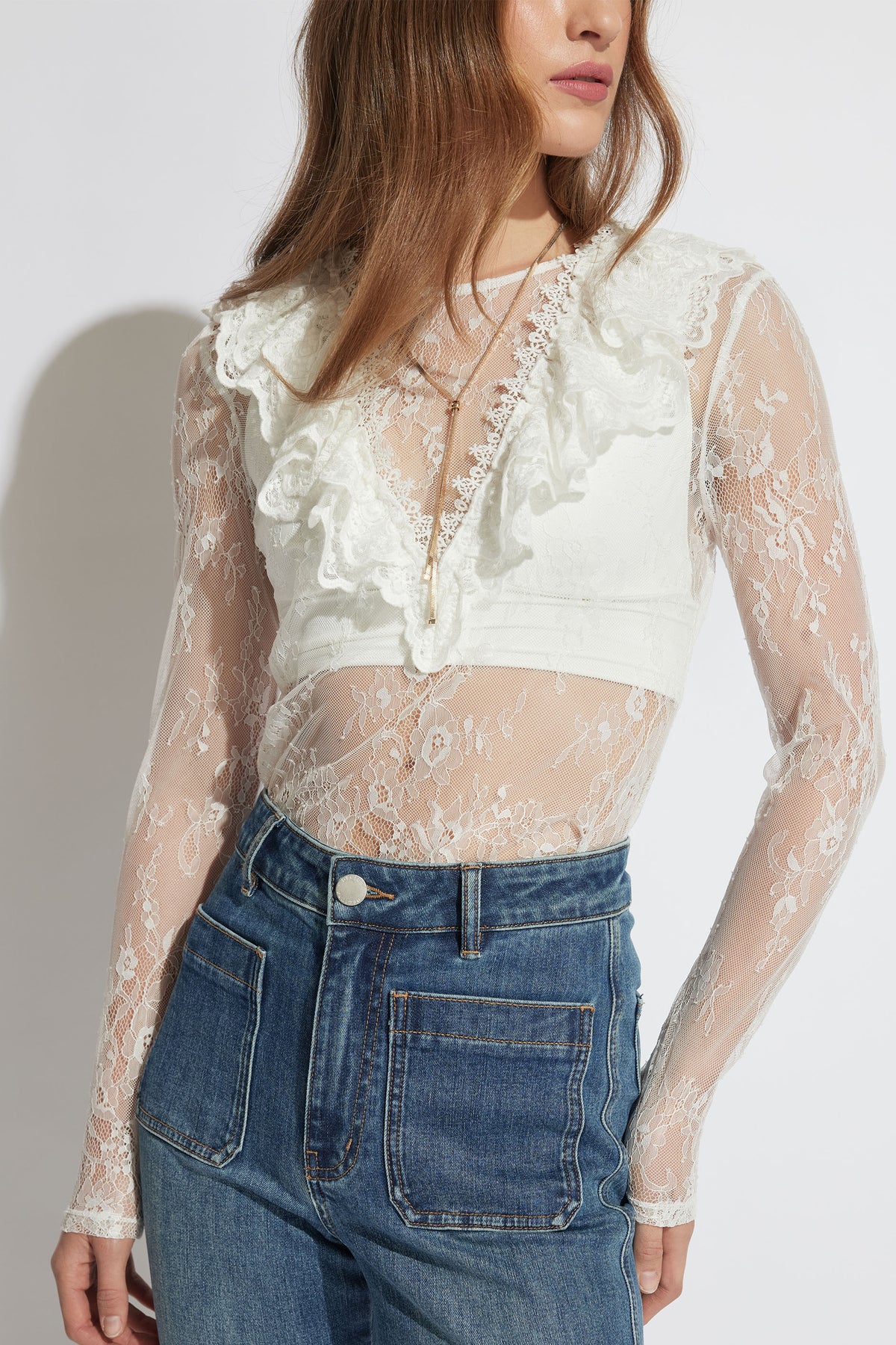 Woman wearing a sheer white lace long-sleeve top with ruffle detailing paired with high-waisted blue denim jeans