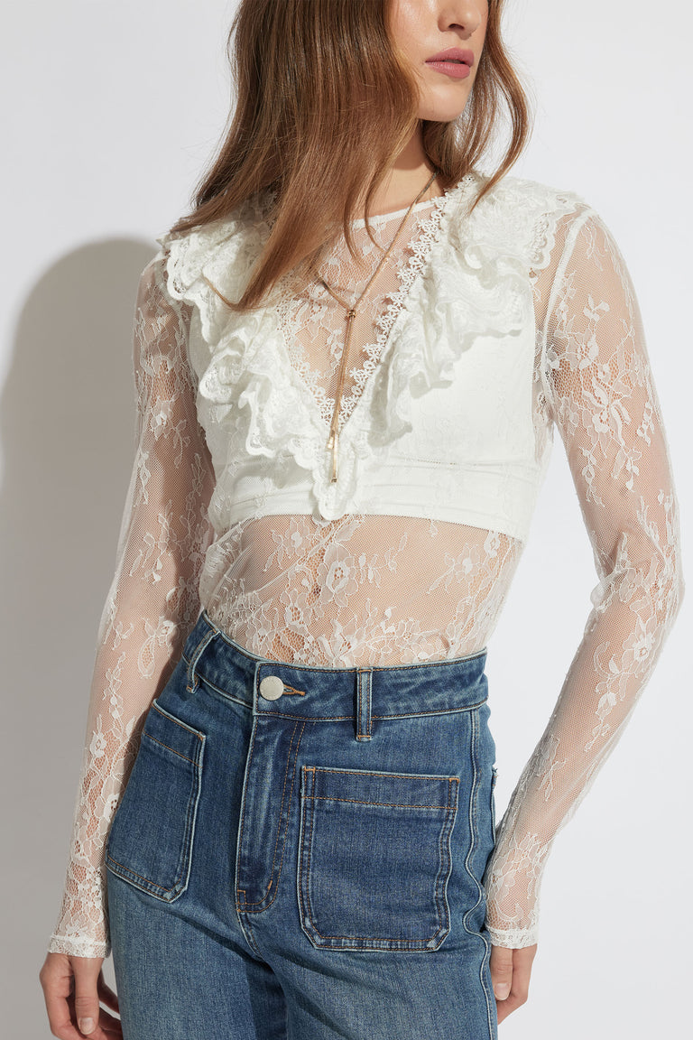 Woman wearing a sheer white lace long-sleeve top with ruffle detailing paired with high-waisted blue denim jeans