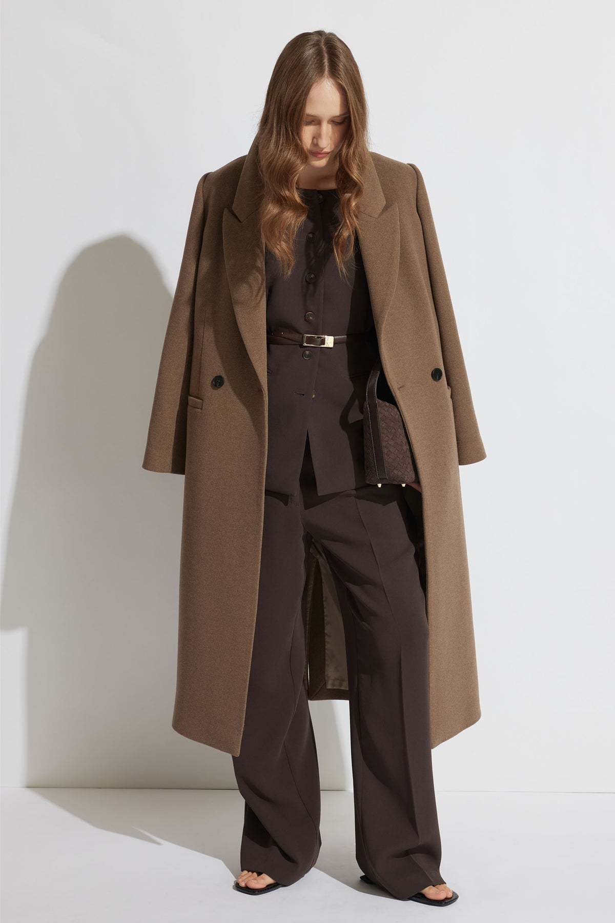 Woman wearing a tailored dark brown vest and wide-leg pants layered under a long camel coat and black sandals