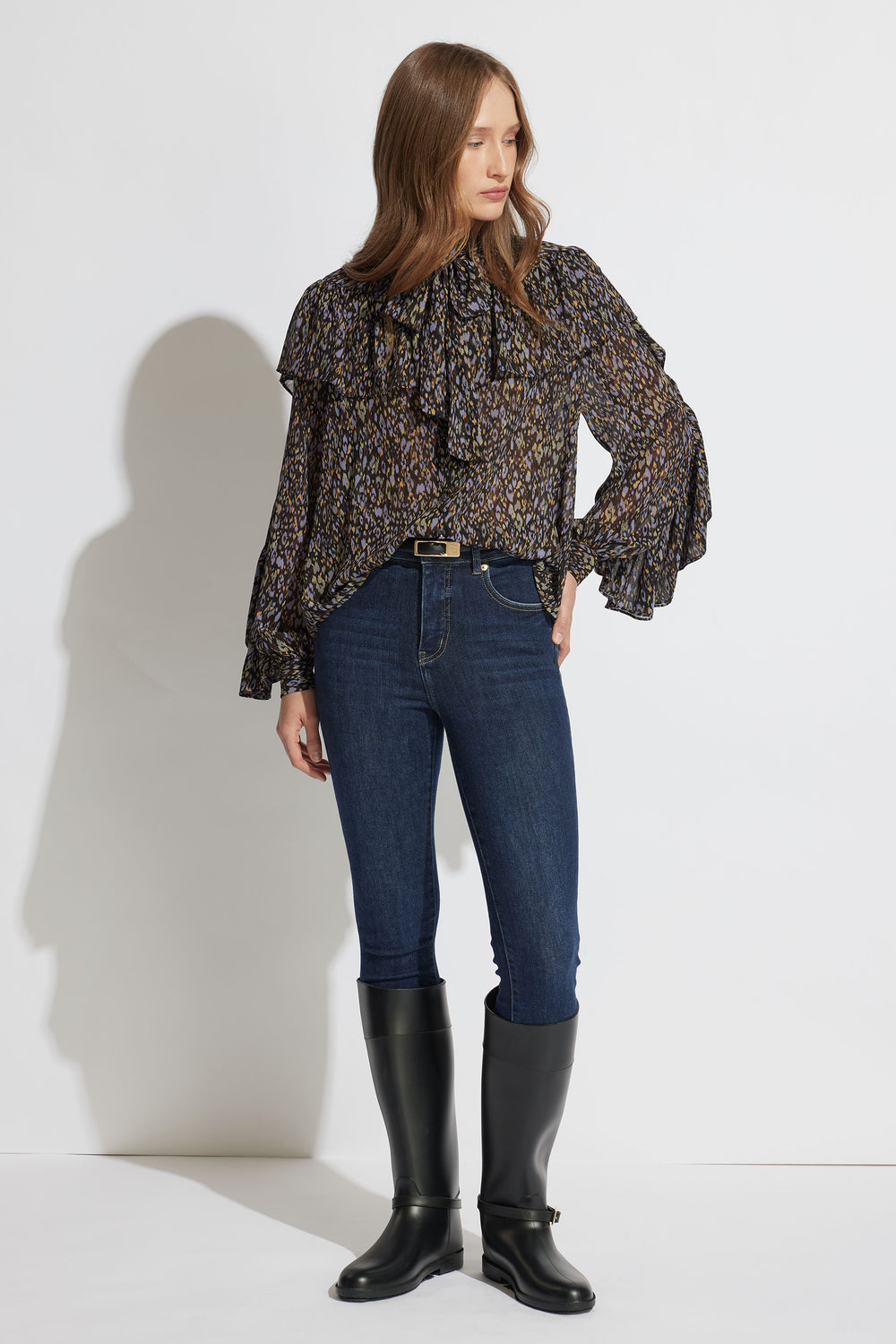 Woman wearing a patterned ruffle blouse with high-waisted dark skinny jeans and black knee-high boots standing against a white wall