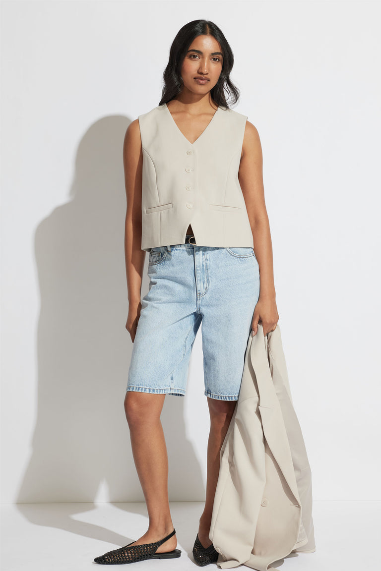 Woman wearing a tailored cream vest with light blue denim long shorts and black woven flats, holding a matching jacket.