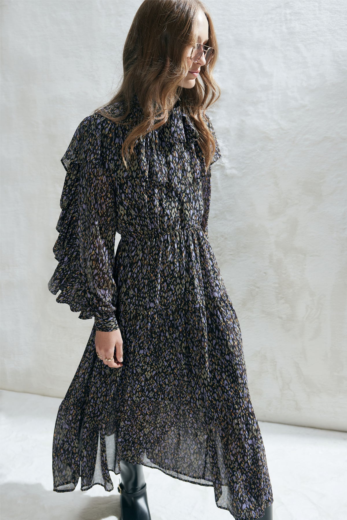 Woman wearing a dark floral midi dress with tiered ruffles, long sleeves, and a tied V neckline, styled with boots.