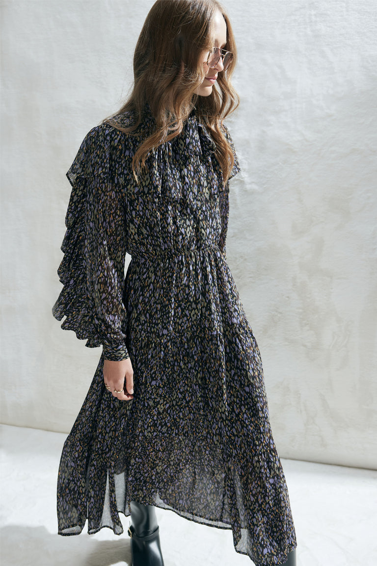 Woman wearing a dark floral midi dress with tiered ruffles, long sleeves, and a tied V neckline, styled with boots.