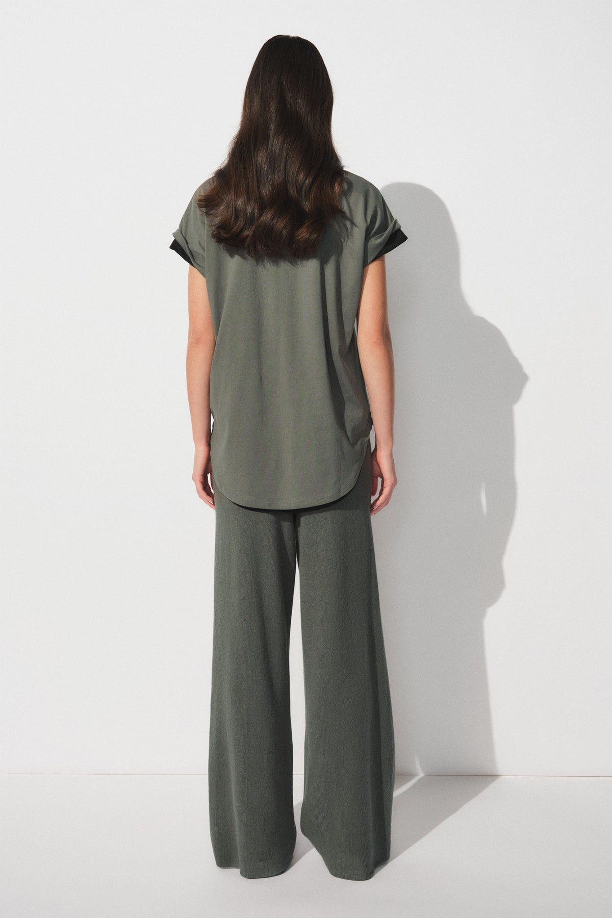 Woman wearing a dark olive tee with a curved hem, viewed from behind