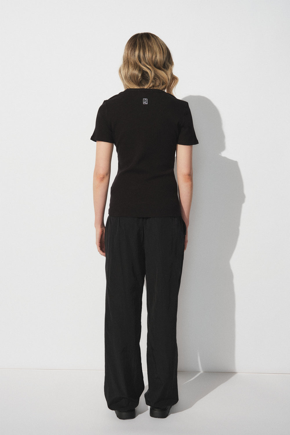 Woman with wavy blonde hair wearing a fitted black short-sleeve top and relaxed black parachute-style joggers, back view.