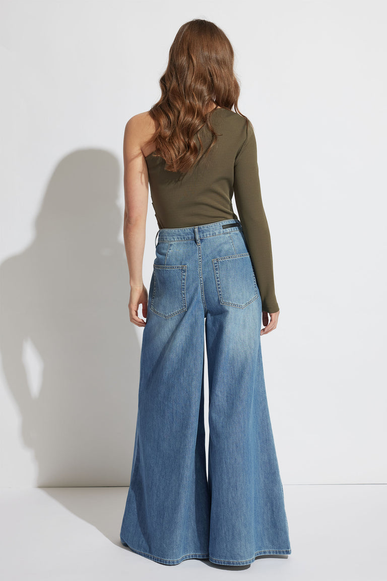 Woman with wavy hair wearing an olive one-shoulder long sleeve top paired with wide-leg light blue jeans