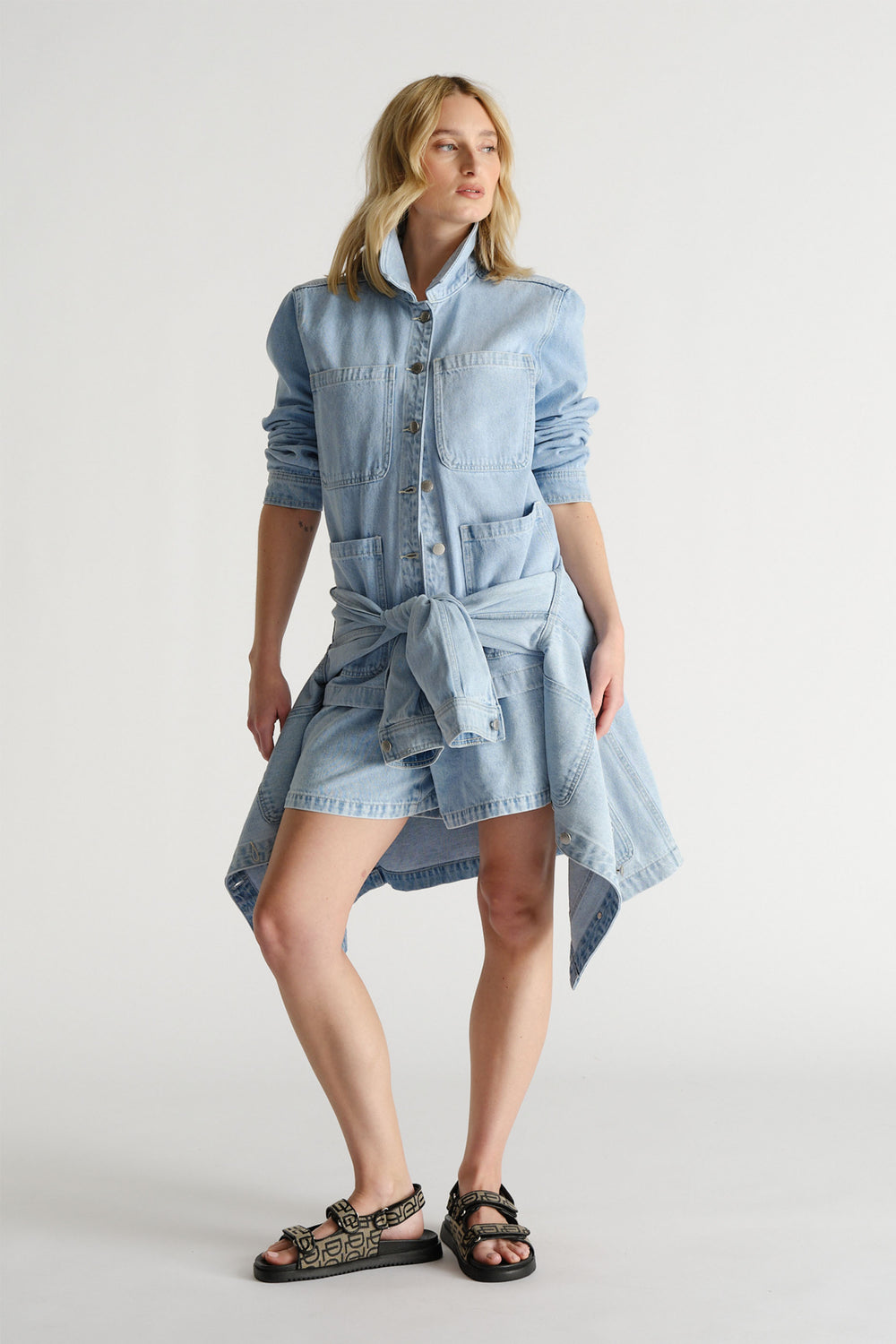 Woman wearing a light blue denim shacket with chest pockets and sleeves pushed up, styled with a tied denim jacket and sandals.