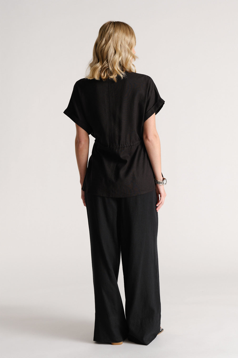 Back view of a woman wearing a black short-sleeve linen blend shirt with relaxed black wide-leg pants.