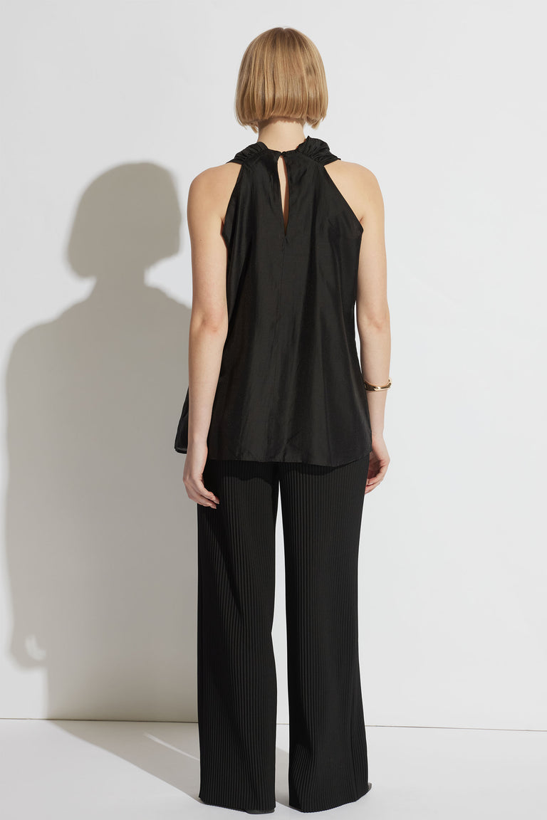 Woman wearing a sleeveless black draped top with a keyhole back and wide-leg black pants against a plain backdrop