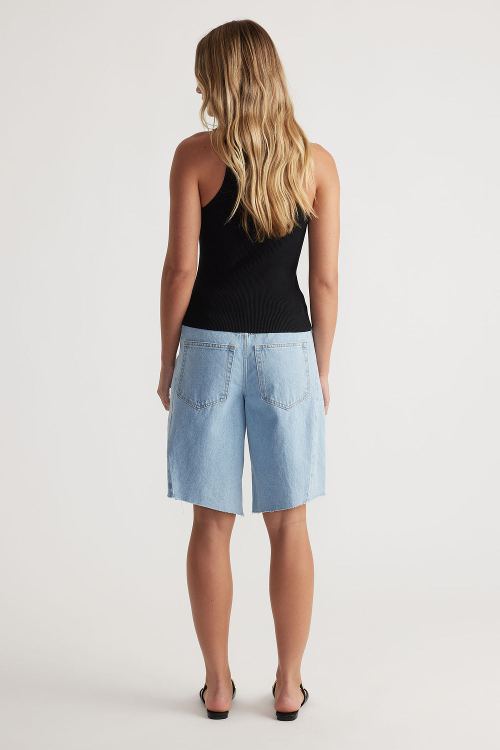 Teen girl shown from behind wearing a black sleeveless top paired with light blue denim jorts and black sandals