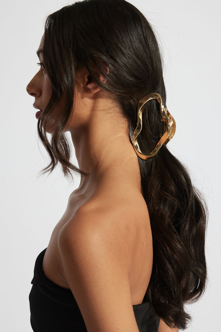 Profile of woman with wavy dark hair styled in low ponytail, secured by a large sculptural gold hair claw accessory.