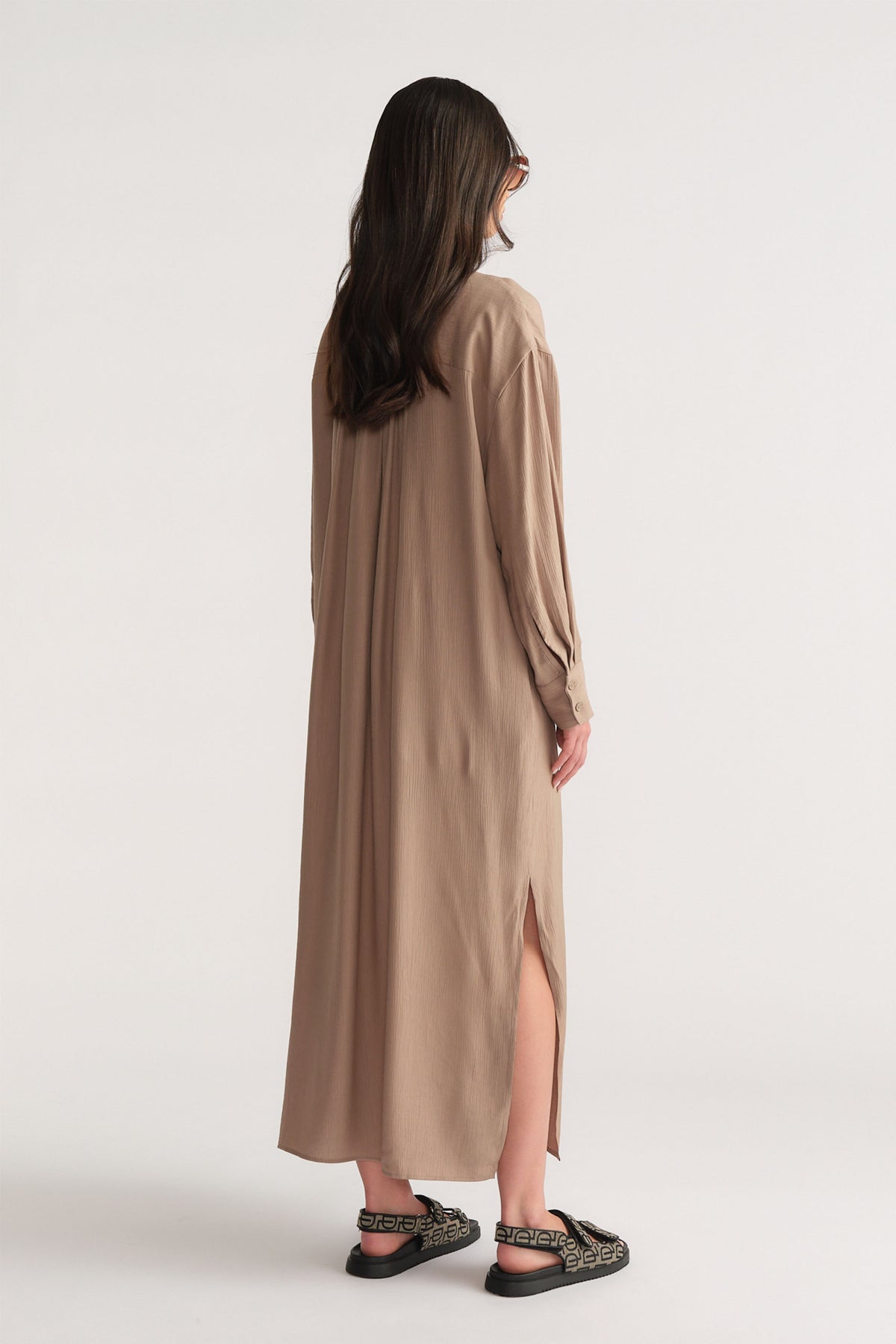 Rear view of a woman wearing a relaxed barley-colored textured shirt dress with side slit and patterned black sandals
