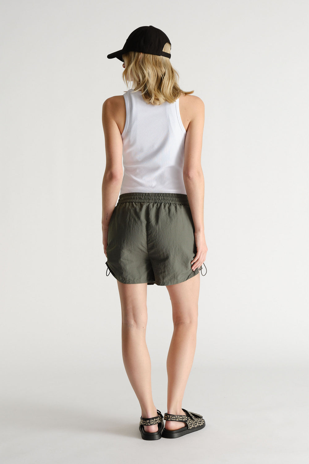 Woman wearing olive green nylon sport shorts with an elastic waistband, white tank top, black cap, and printed sandals.