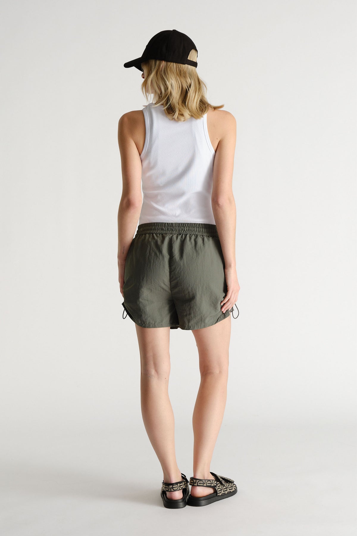 Woman wearing olive green nylon sport shorts with an elastic waistband, white tank top, black cap, and printed sandals.