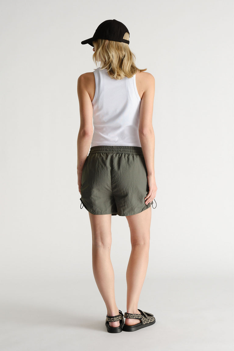 Woman wearing olive green nylon sport shorts with an elastic waistband, white tank top, black cap, and printed sandals.
