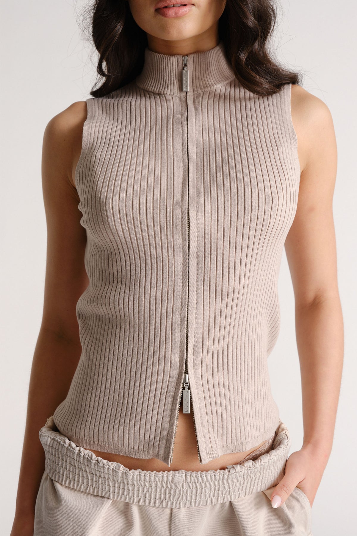 Sleeveless beige ribbed knit top with a high neck and front zipper, styled with relaxed waist pants.