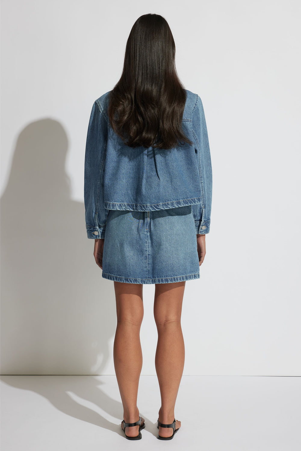 Woman seen from behind wearing a relaxed fit light blue denim jacket and matching denim skirt with black sandals