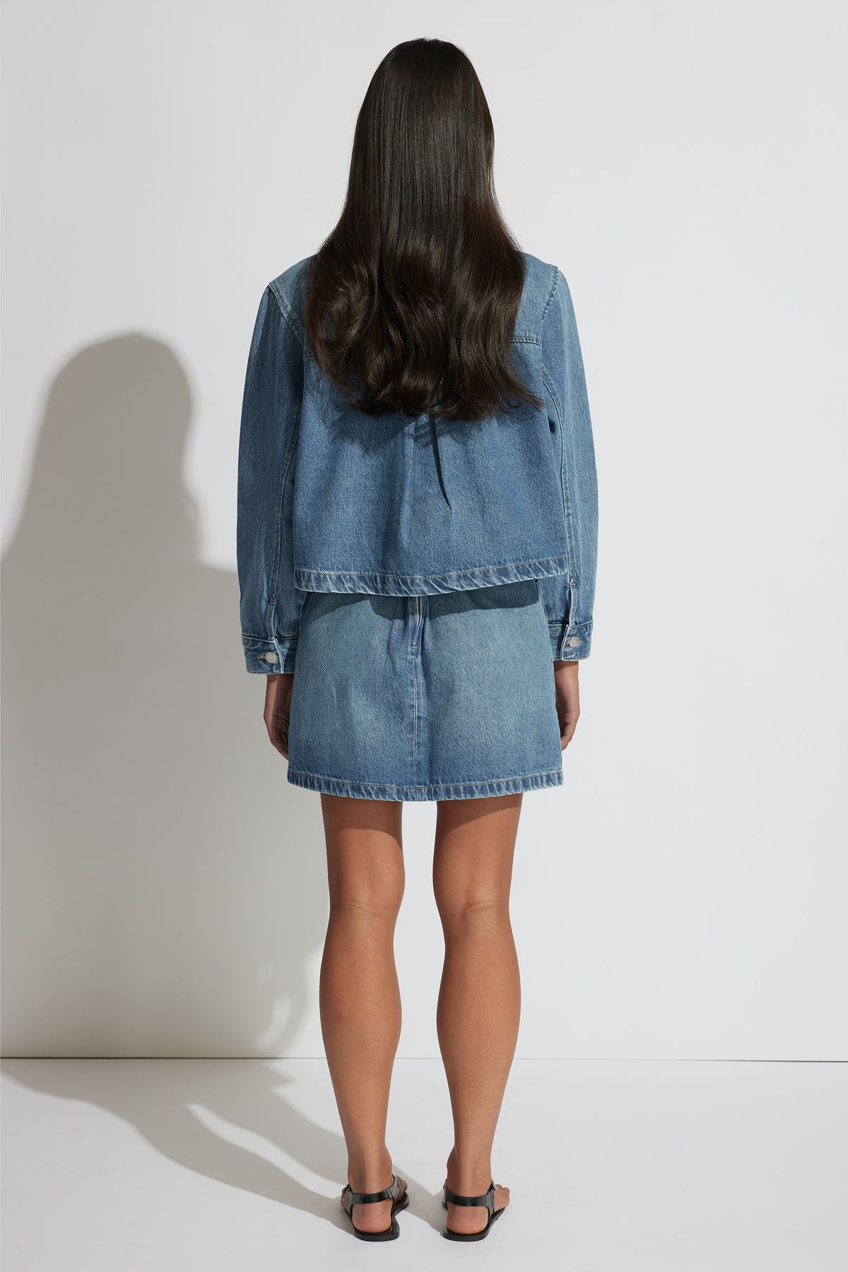 Woman seen from behind wearing a relaxed fit light blue denim jacket and matching denim skirt with black sandals