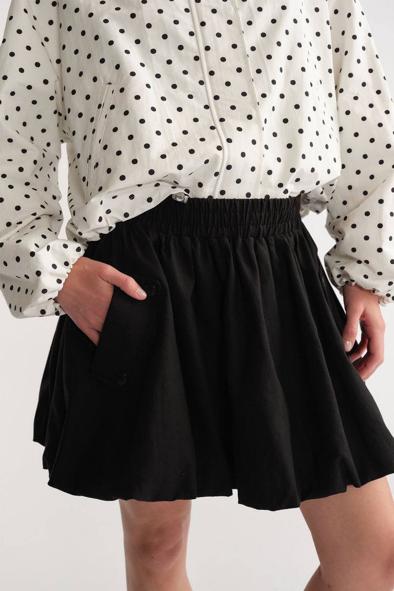 Woman wearing a black bubble mini skirt with elastic waist and side button pocket paired with a white polka dot blouse