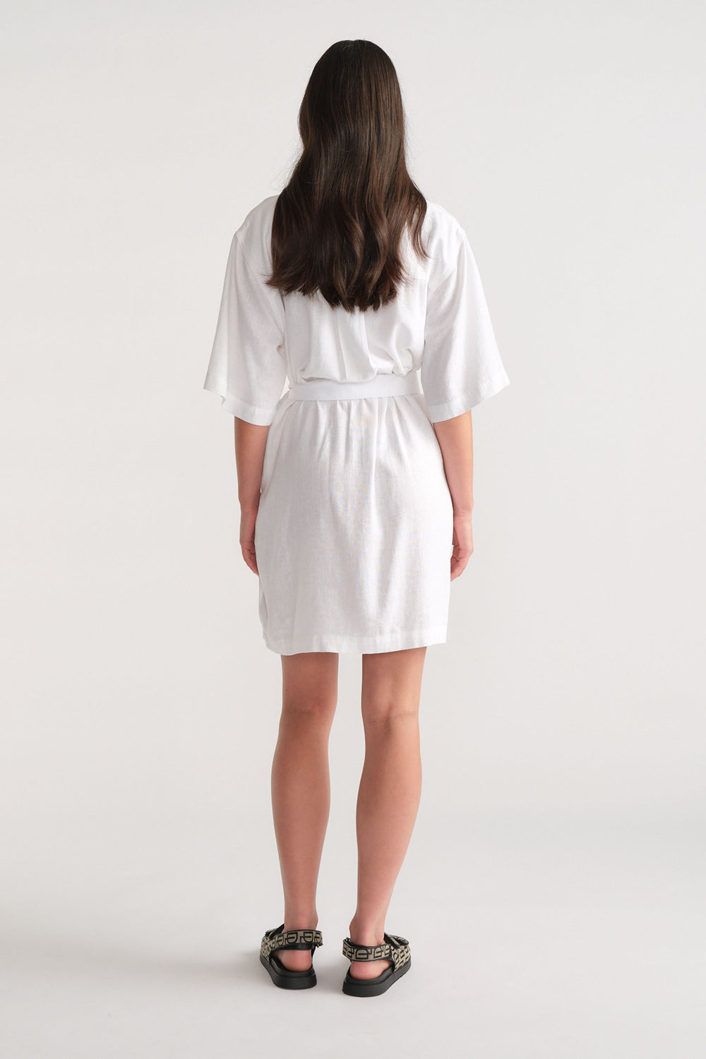 Woman wearing a white short linen blend dress with loose sleeves, styled with patterned black sandals, shown from the back.