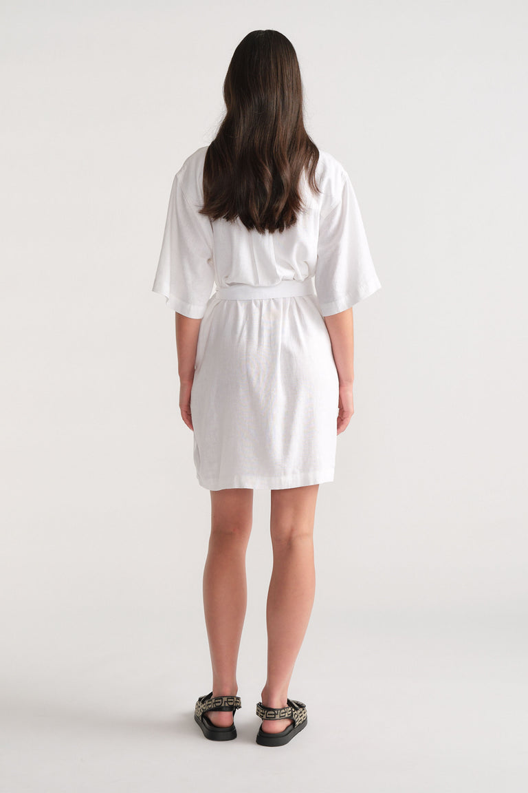 Woman wearing a white short linen blend dress with loose sleeves, styled with patterned black sandals, shown from the back.
