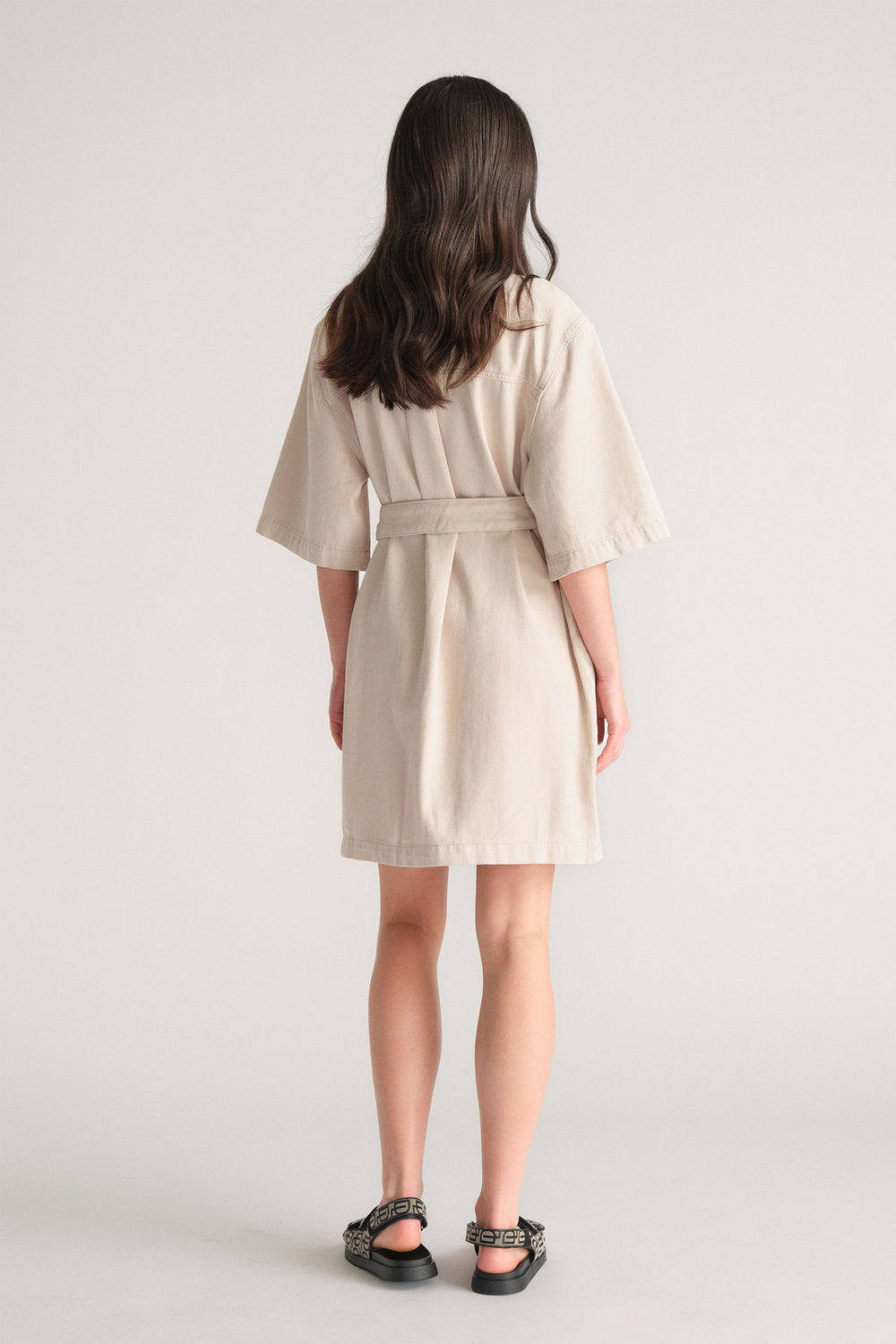 Back view of model wearing beige D-Luxe Denim Short Dress with elbow sleeves and optional waist tie, gallery image 3 in portrait.