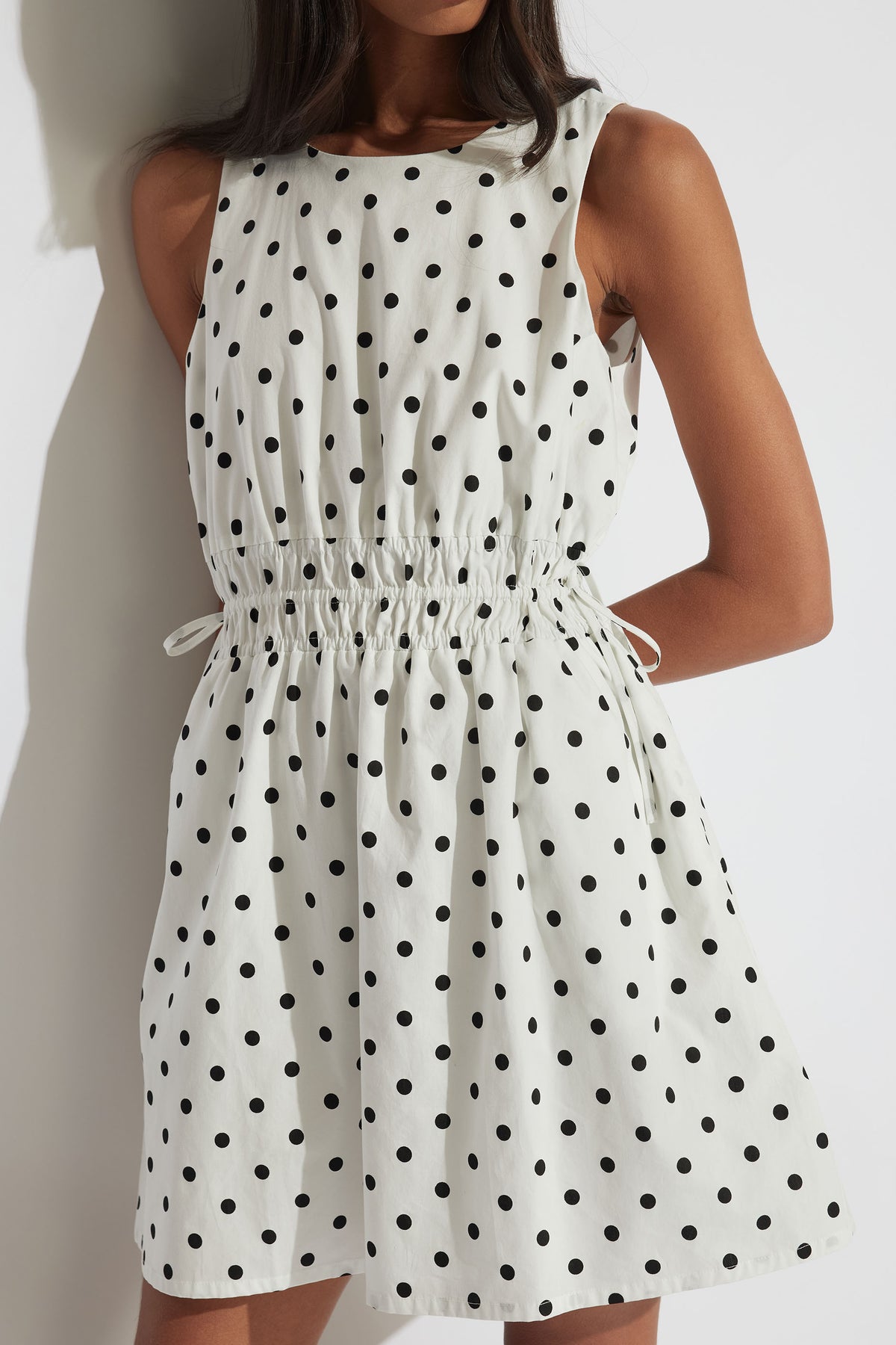 Cali Spot Mini Dress in portrait, featuring sleeveless design and shirred waist with adjustable ties in polka dot print.