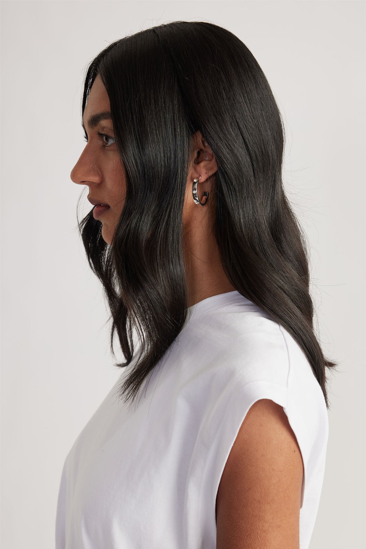 Woman with sleek dark hair wearing silver hoop earrings and a white sleeveless top against a neutral background