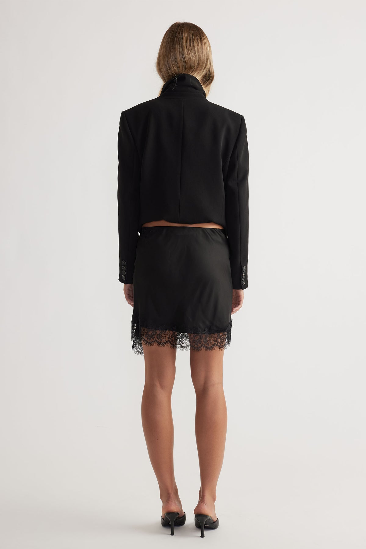 Back view of model wearing Karla Lace Mini Skirt with lace trim, paired with cropped black jacket, gallery position 3 portrait.