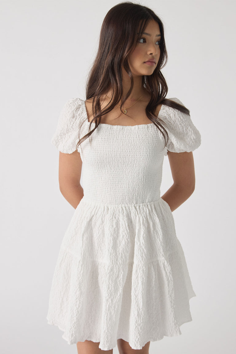 Girl wearing a white textured mini dress with puff sleeves and a smocked bodice, looking to the side.