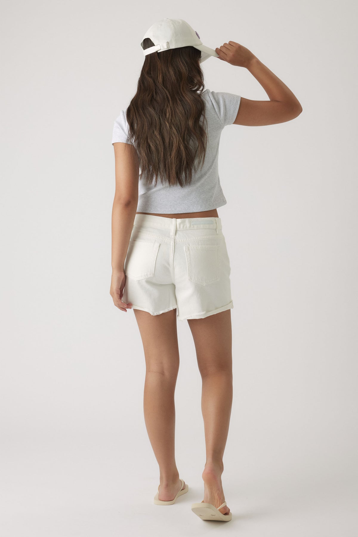 Girl wearing white denim shorts with raw hem paired with a grey crop top and white cap, casual summer look