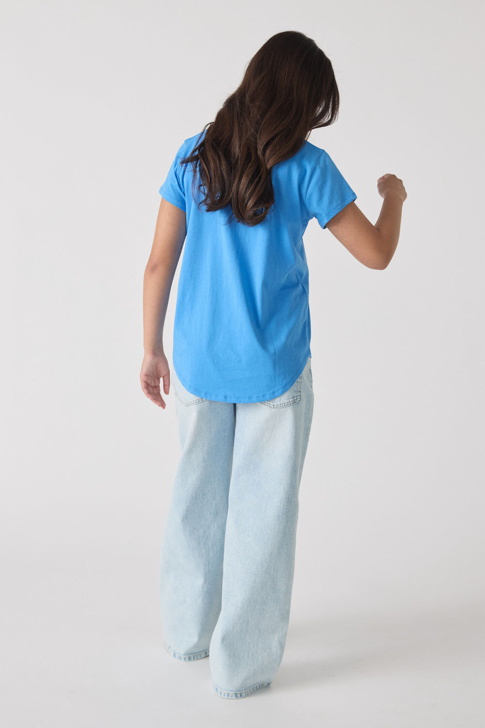 Girl wearing a vibrant blue tee with short sleeves and relaxed light wash wide-leg jeans, shown from behind.